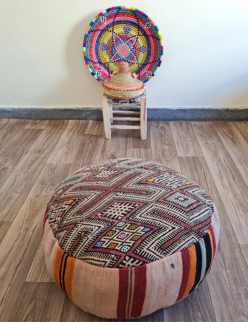 A vintage Kilim floor cushion on a wood floor. In the background is a small wooden stool with a colorful Moroccan basket and plate.