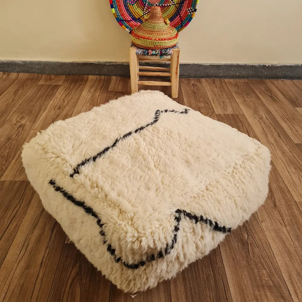 A square Beni Ourain floor cushion with abstract black lines on a wood floor. In the background, a small wooden stool displays a colorful Moroccan woven basket