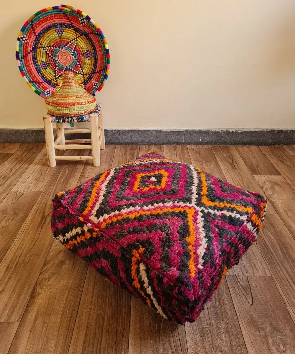 A square vintage Boucherouite floor cushion in vibrant magenta and orange on a wood floor. In the background, a small wooden stool displays a colorful Moroccan woven basket