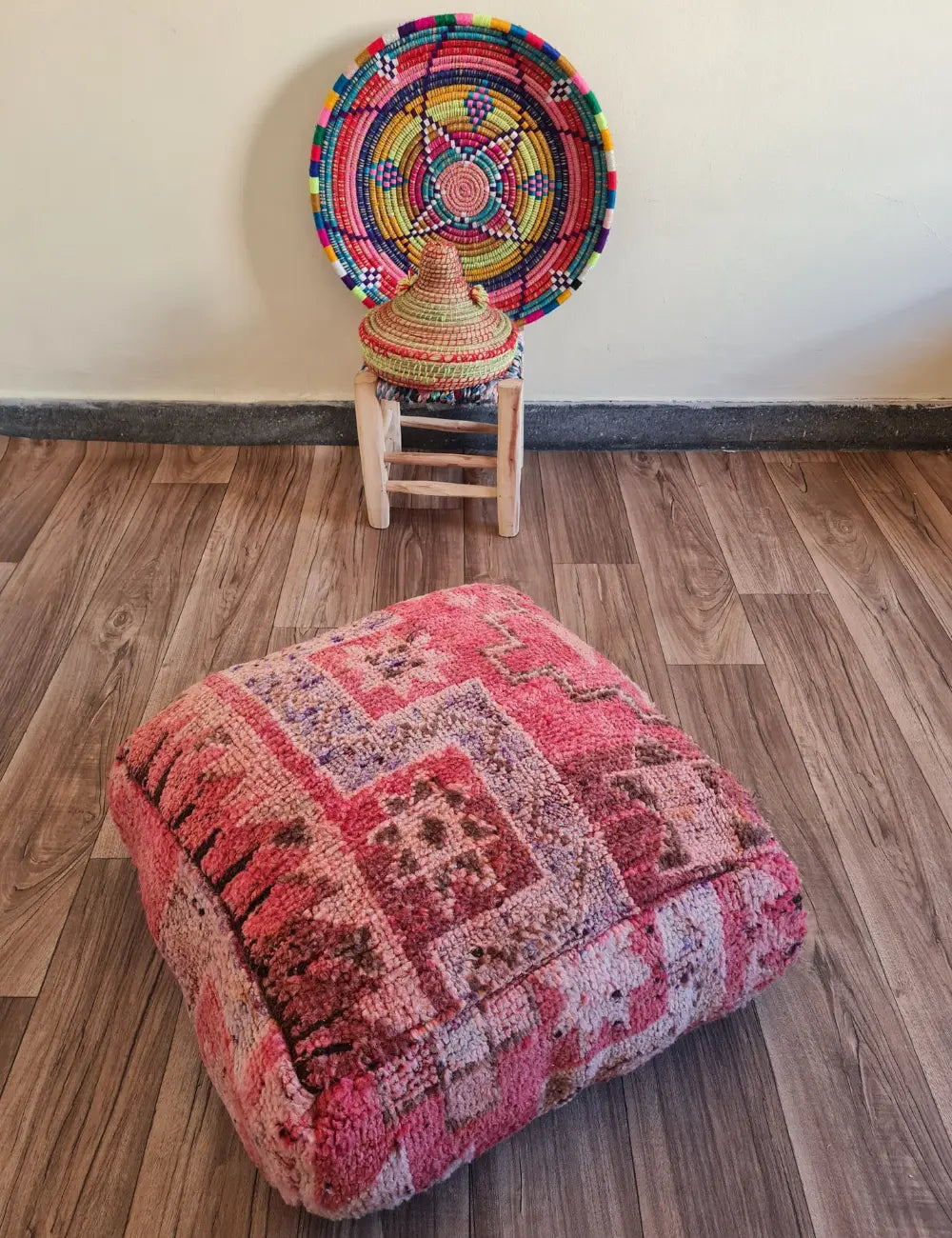 A square vintage rug floor cushion in shades of pink on a wood floor. In the background, a small wooden stool displays a colorful Moroccan woven basket