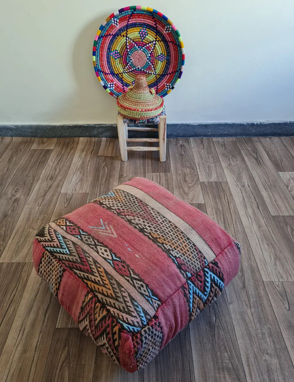 A square vintage Kilim floor cushion in shades of dusty rose on a wood floor. In the background, a small wooden stool displays a colorful Moroccan woven basket