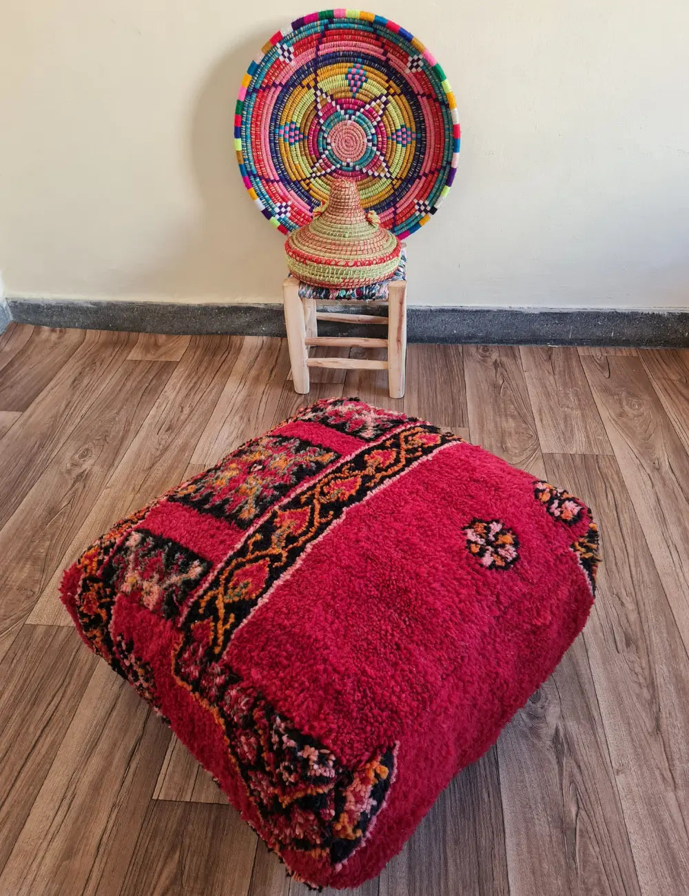 A square vintage rug floor cushion in deep red on a wood floor. In the background, a small wooden stool displays a colorful Moroccan woven basket