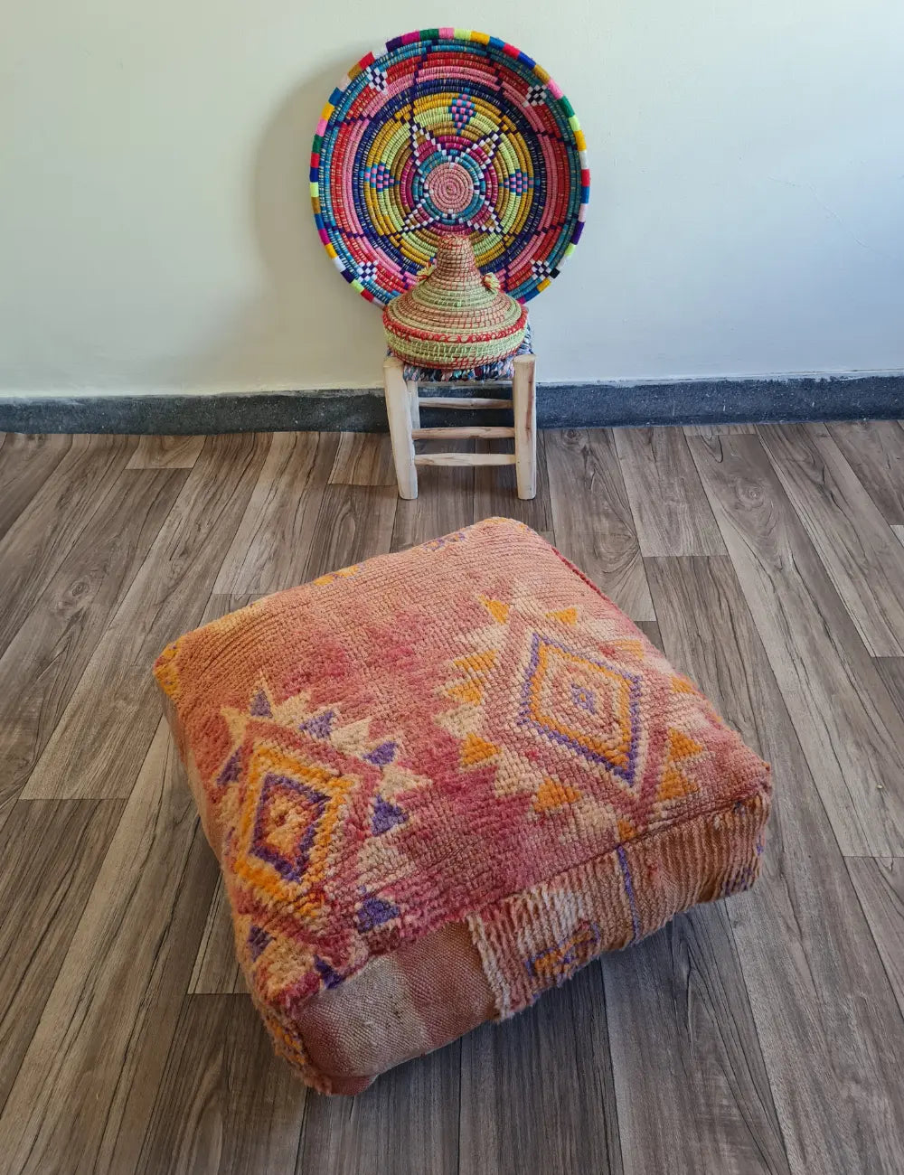 A square vintage rug floor cushion in sun-faded reds on a wood floor. In the background, a small wooden stool displays a colorful Moroccan woven basket