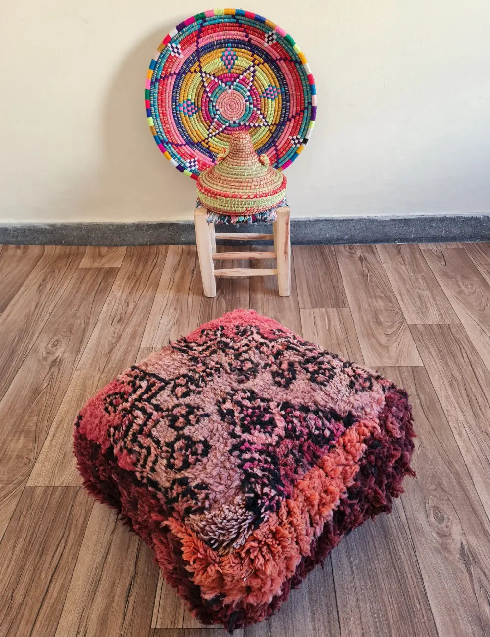 A square vintage rug floor cushion in shades of deep pink and red on a wood floor. In the background, a small wooden stool displays a colorful Moroccan woven basket
