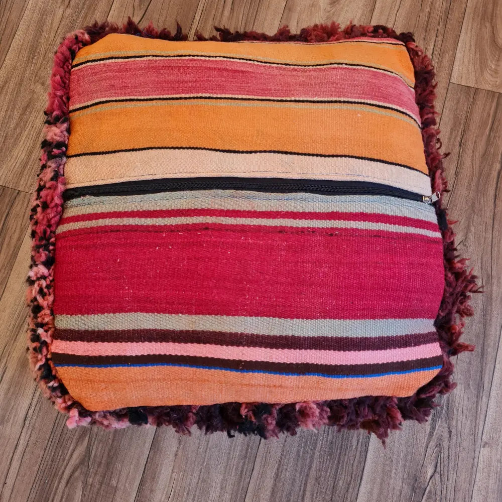 The bottom of the square pouf, showing a flat-woven striped Kilim fabric in shades of red, pink, orange, and green, with a large black zipper for stuffing