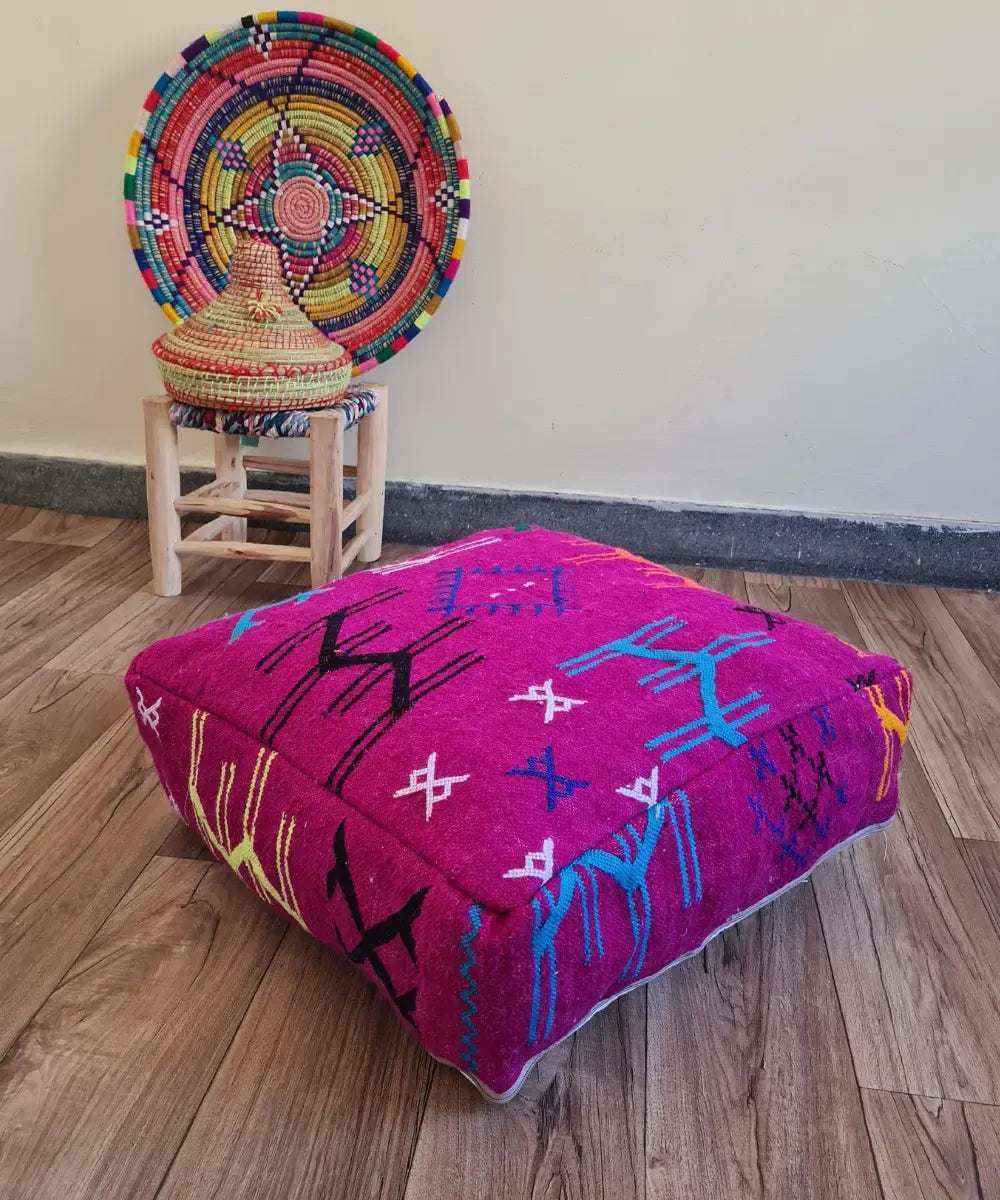 A square modern floor cushion with fuchsia embroidery on a wood floor. In the background, a small wooden stool displays a colorful Moroccan woven basket