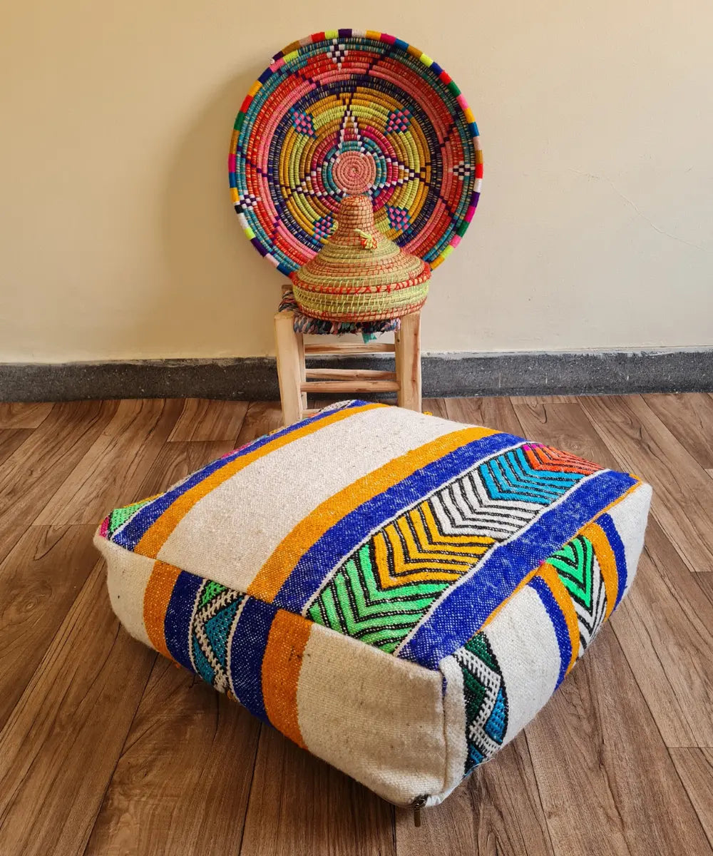 A square modern floor cushion with vibrant geometric patterns on a wood floor. In the background, a small wooden stool displays a colorful Moroccan woven basket