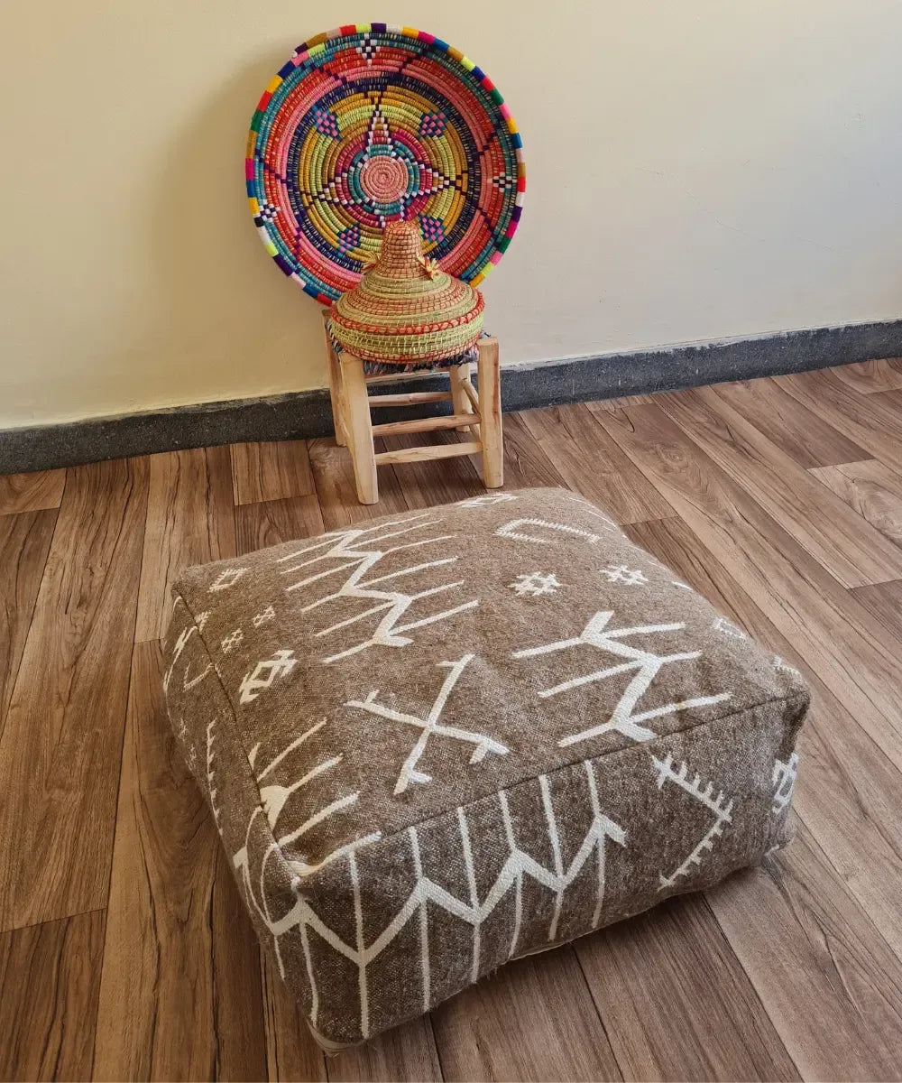 A square modern floor cushion with black and white tribal patterns on a wood floor. In the background, a small wooden stool displays a colorful Moroccan woven basket