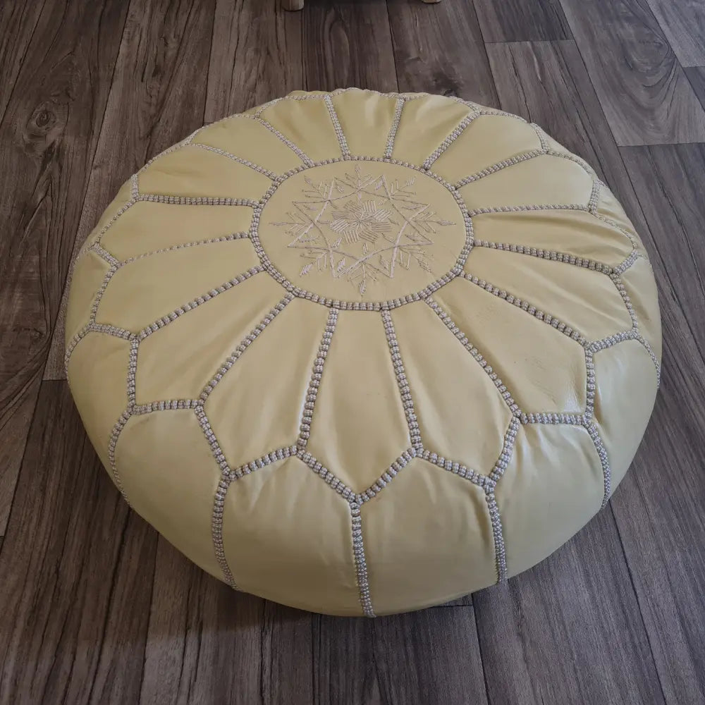 An angled, three-quarter view of a beige leather Moroccan pouf, highlighting the intricate tonal stitching and embroidery