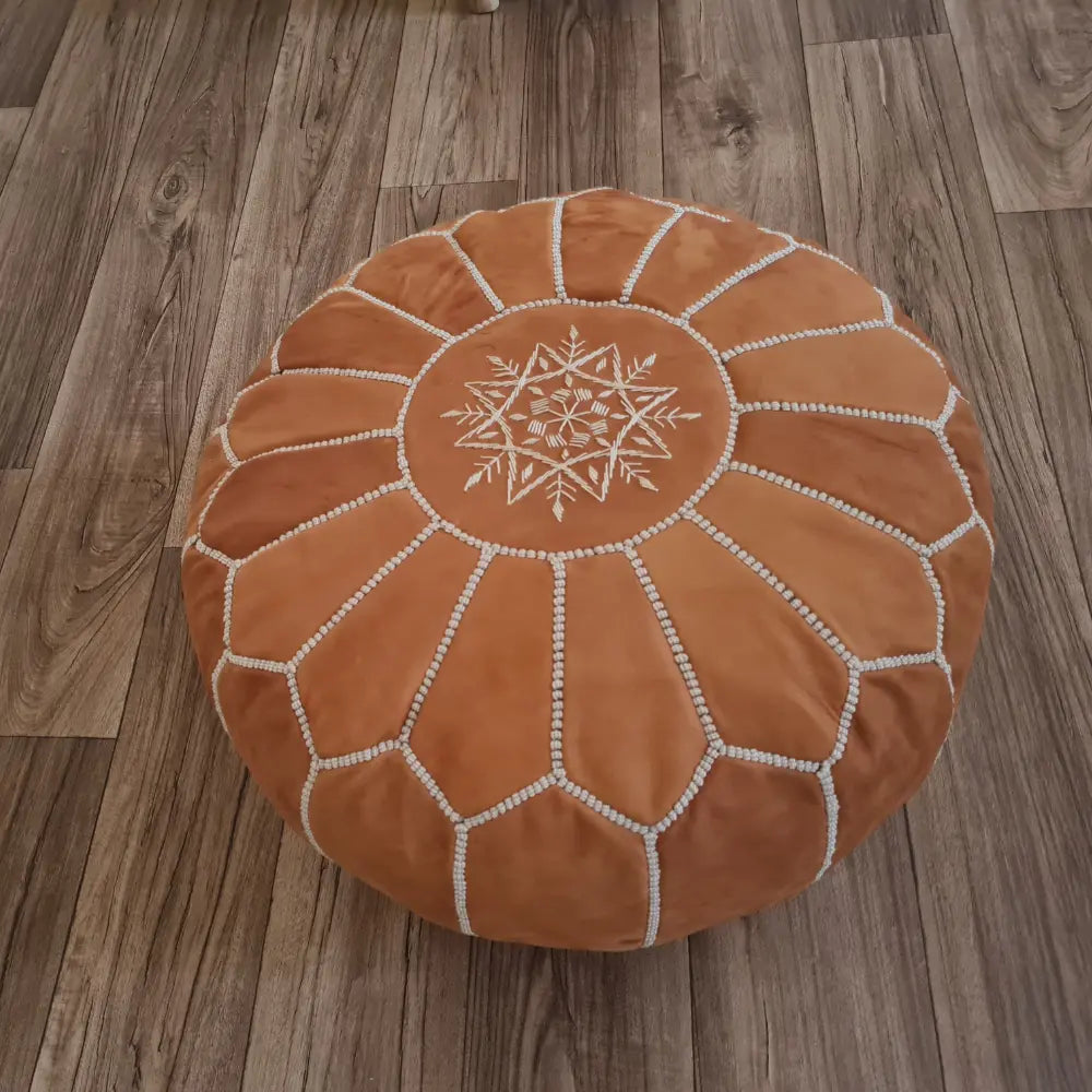 An angled, three-quarter view of a tan suede Moroccan pouf, highlighting the intricate white stitching, embroidery, and soft matte texture