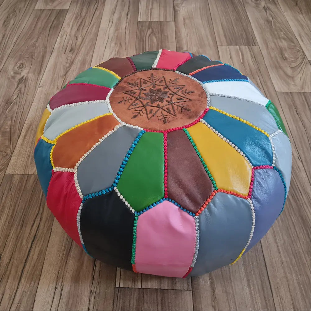 A patchwork Moroccan leather pouf on a wood floor. In the background is a small wooden stool with a colorful Moroccan basket and plate