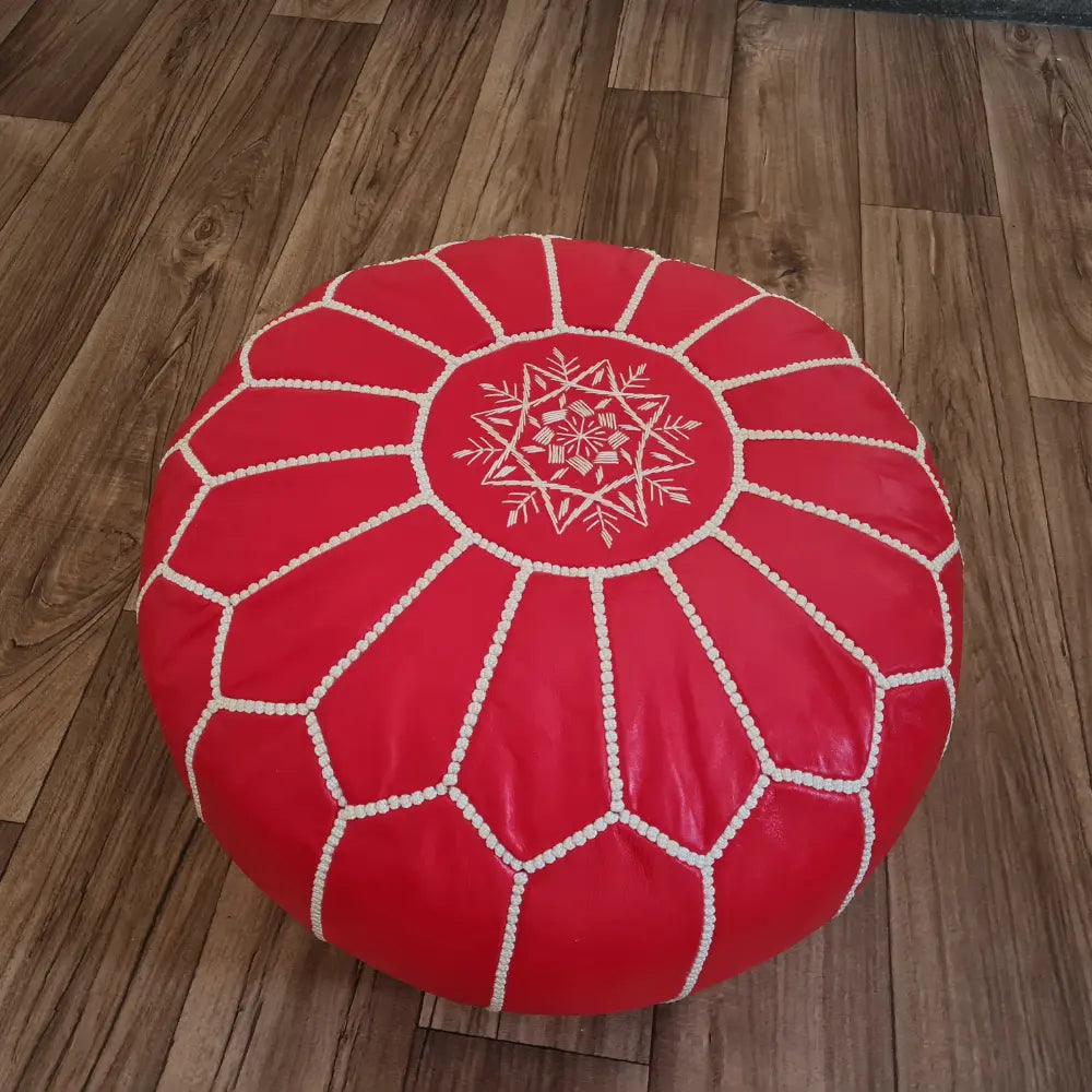 A red Moroccan leather pouf on a wood floor. In the background is a small wooden stool with a colorful Moroccan basket and plate.