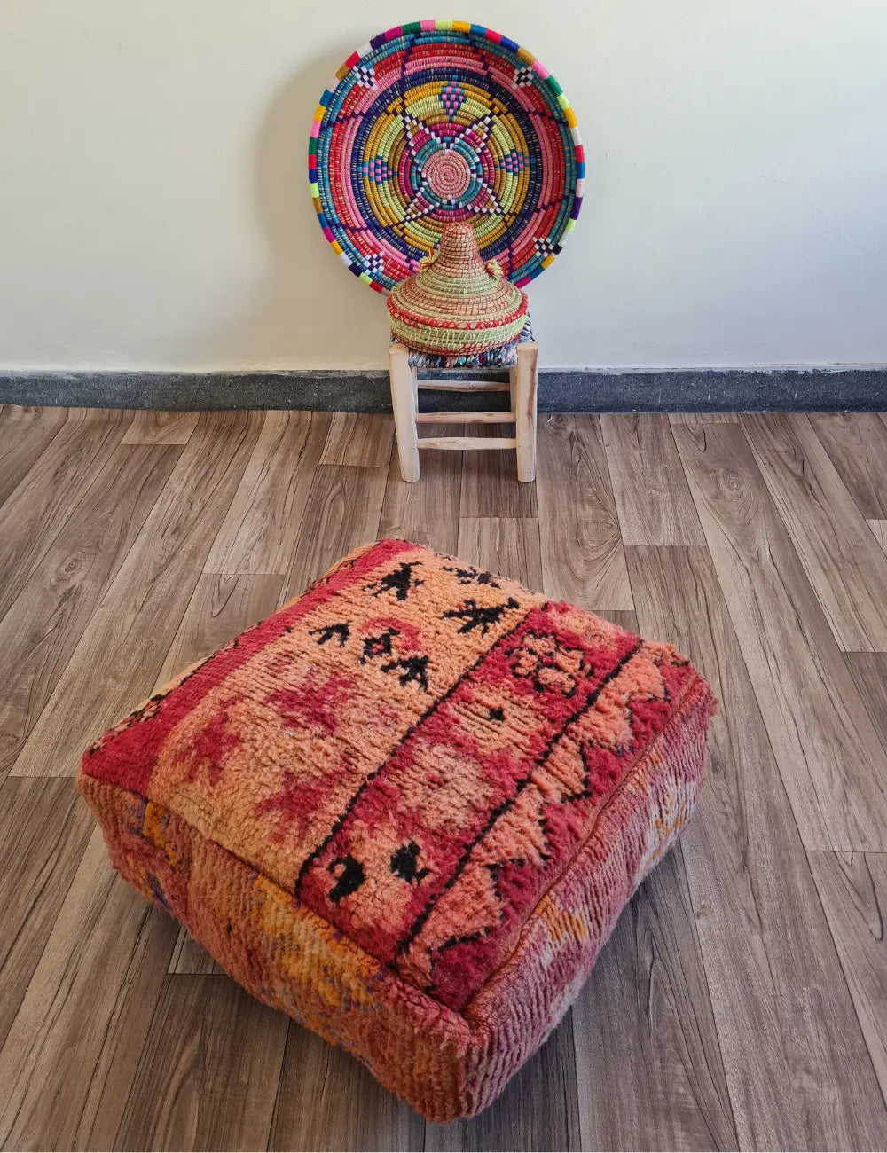 A square vintage rug floor cushion on a wood floor. In the background is a small wooden stool with a colorful Moroccan basket and plate