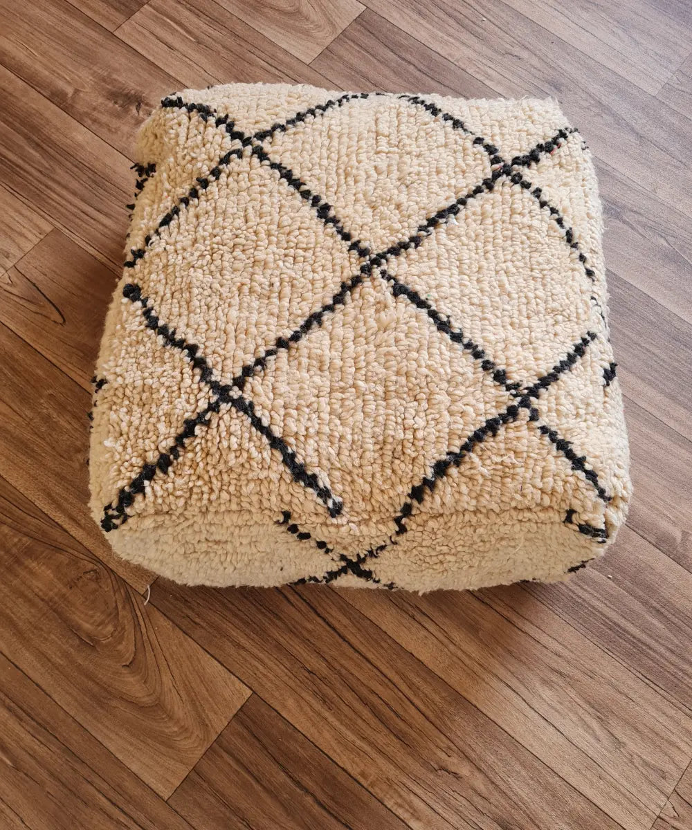 A top-down view of a square, handmade pouf made from a vintage Beni Ourain rug. It features a high-pile shag texture in creamy ivory wool, with a classic black diamond lattice pattern