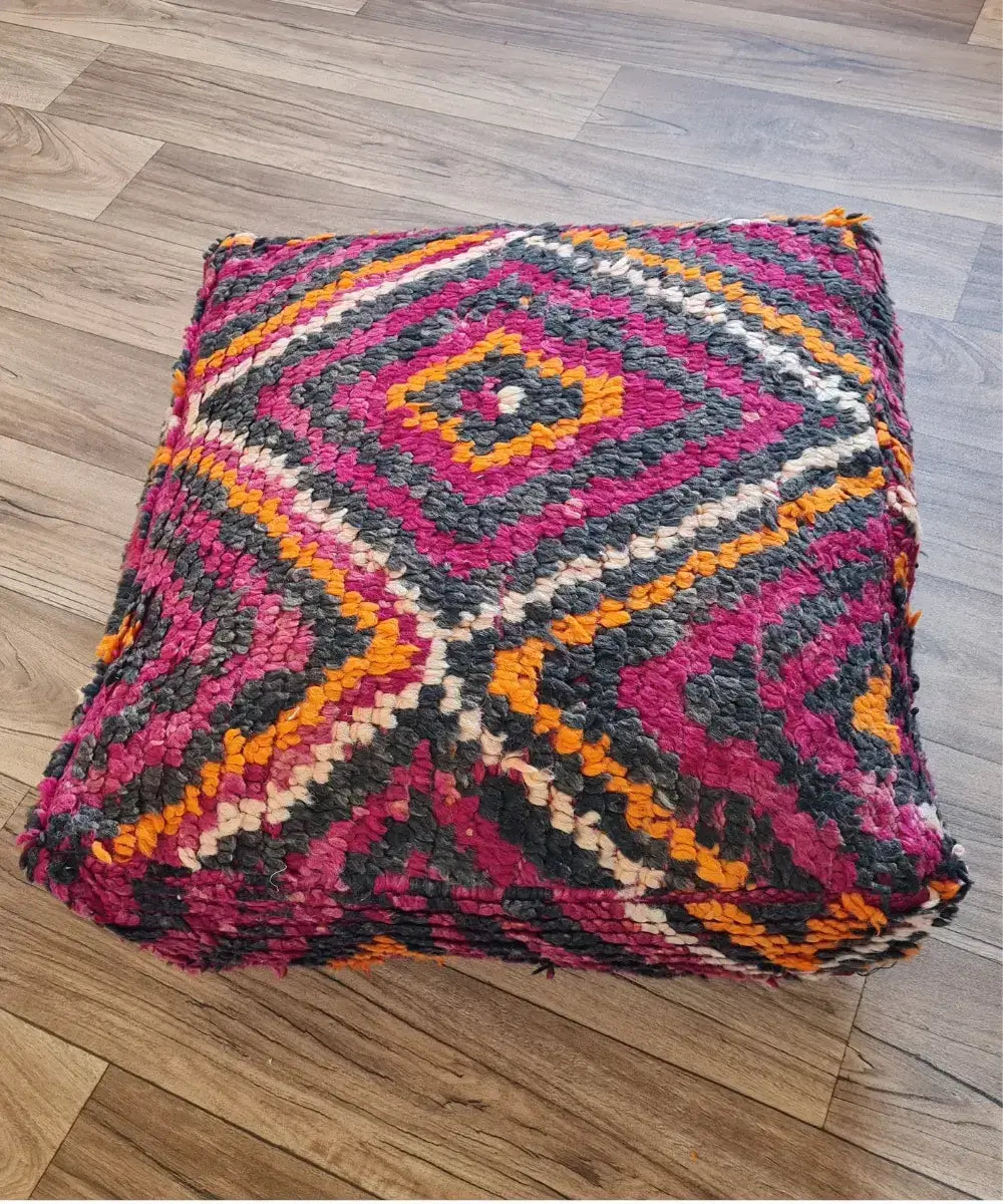 A top-down view of a square, handmade floor cushion made from a vintage Kilim rag rug. It has a high-pile shag texture with a geometric diamond pattern in magenta, orange, charcoal, and white