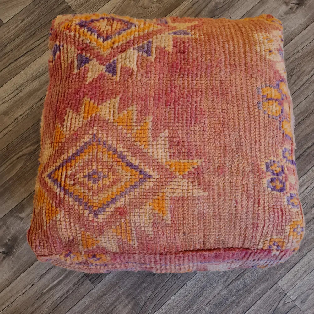 A top-down view of a square, handmade floor cushion made from a vintage Moroccan wool rug. It has a low-pile texture with sun-faded tribal patterns in shades of dusty rose, orange, and purple