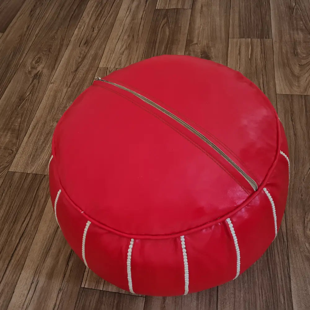 The underside of a red leather pouf, showing the large zipper used for stuffing, set against a dark wood floor