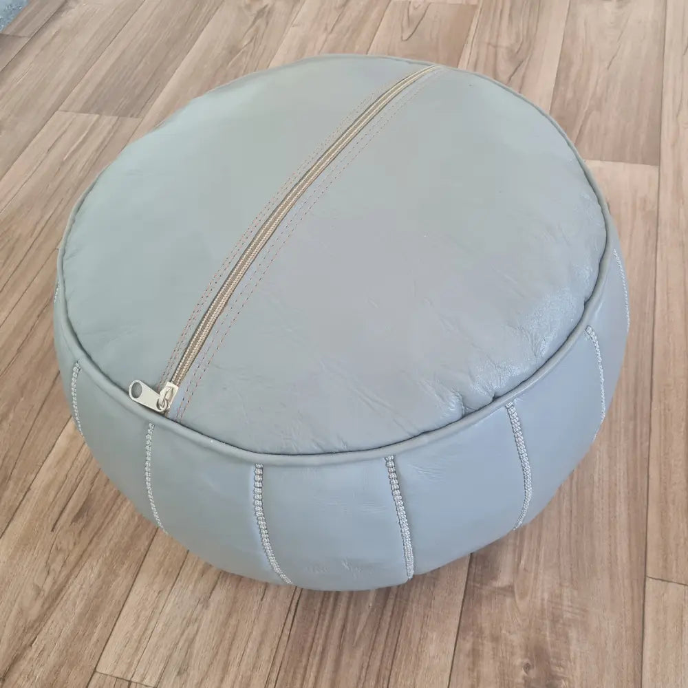 The underside of a grey leather pouf, showing the large zipper used for stuffing, set against a dark wood floor
