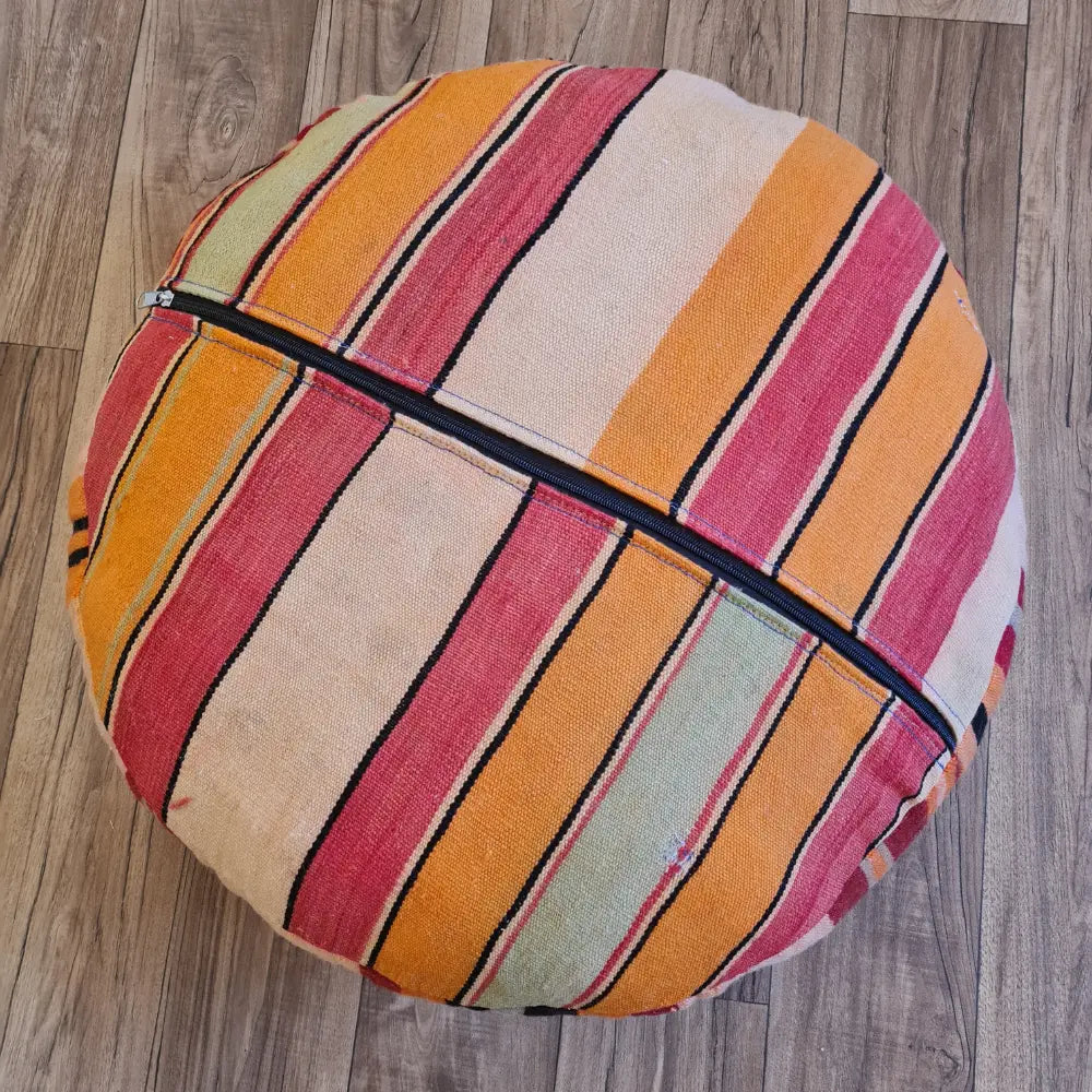The bottom of a round Kilim pouf, showing a striped pattern in pink, orange, and cream, with a large black zipper for stuffing