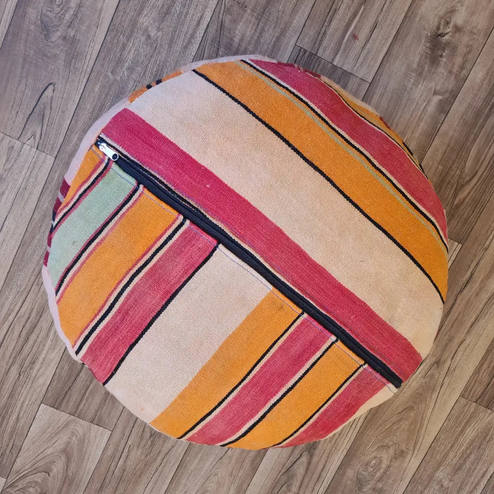 The bottom of a round Kilim pouf, showing a striped pattern in pink, orange, and cream, with a large black zipper for stuffing