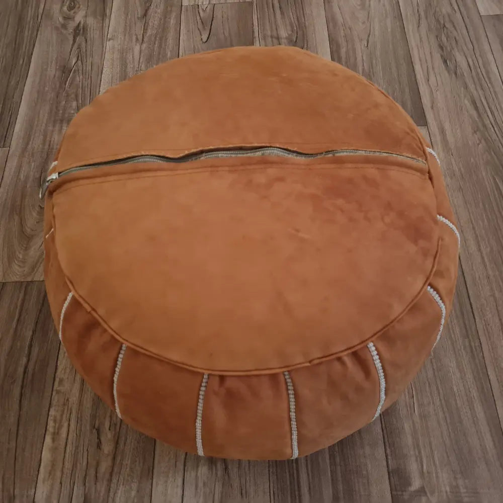 The underside of a tan suede pouf, showing the large zipper used for stuffing, set against a dark wood floor