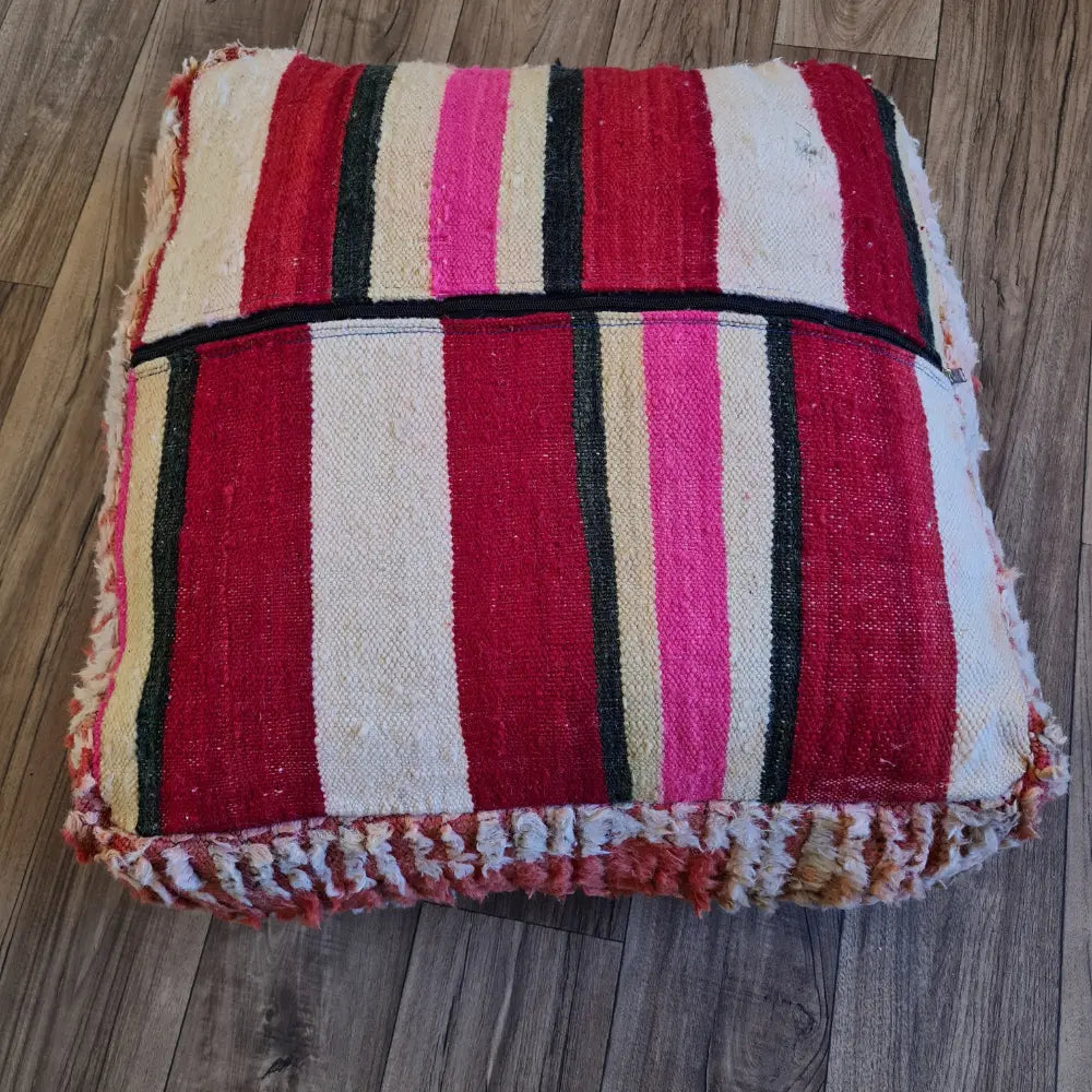 The bottom of a square Boucherouite pouf, showing a striped flat-weave pattern in shades of pink, red, and cream, with a large black zipper for stuffing