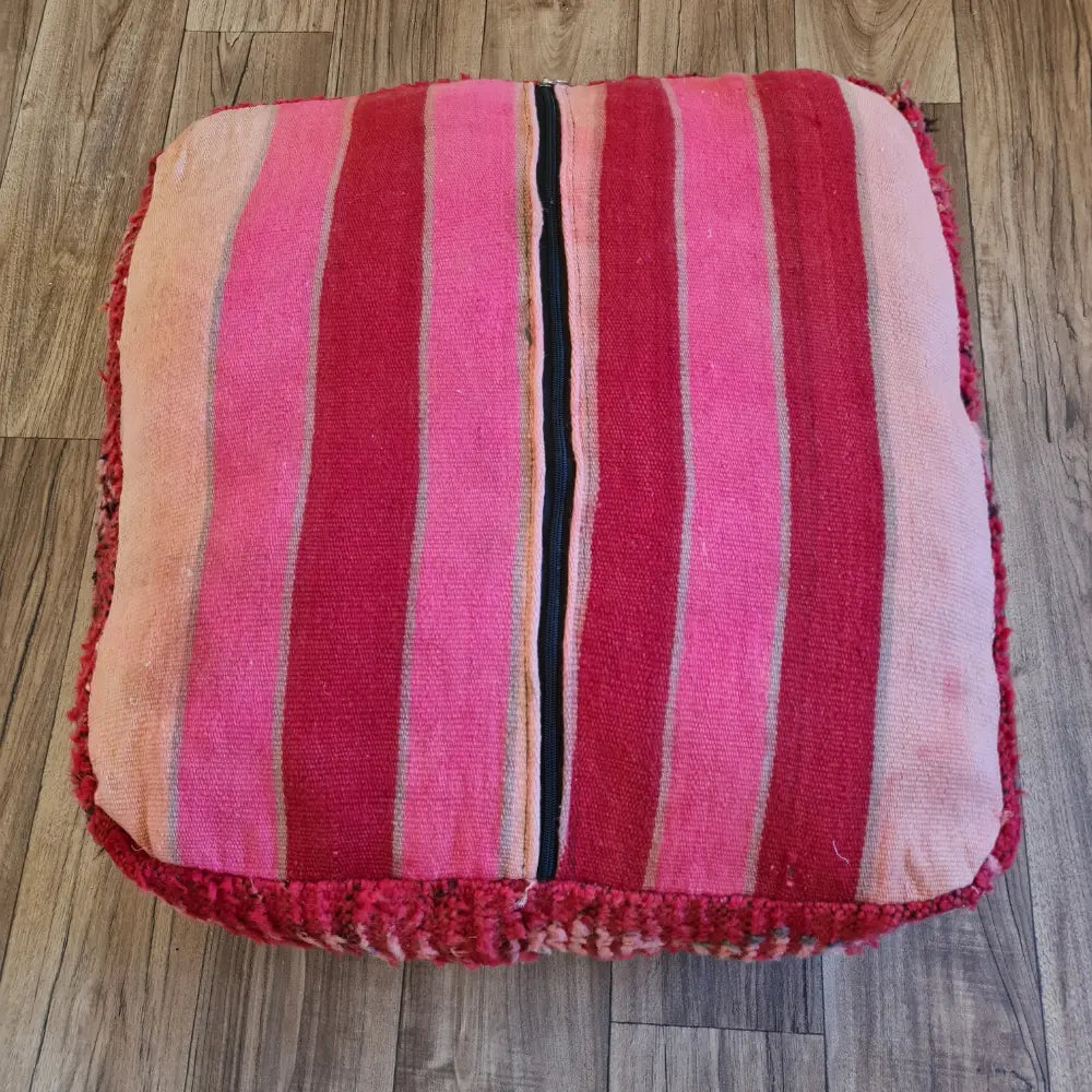 The bottom of a square Boucherouite pouf, showing a striped Kilim pattern in shades of pink and red, with a large black zipper for stuffing
