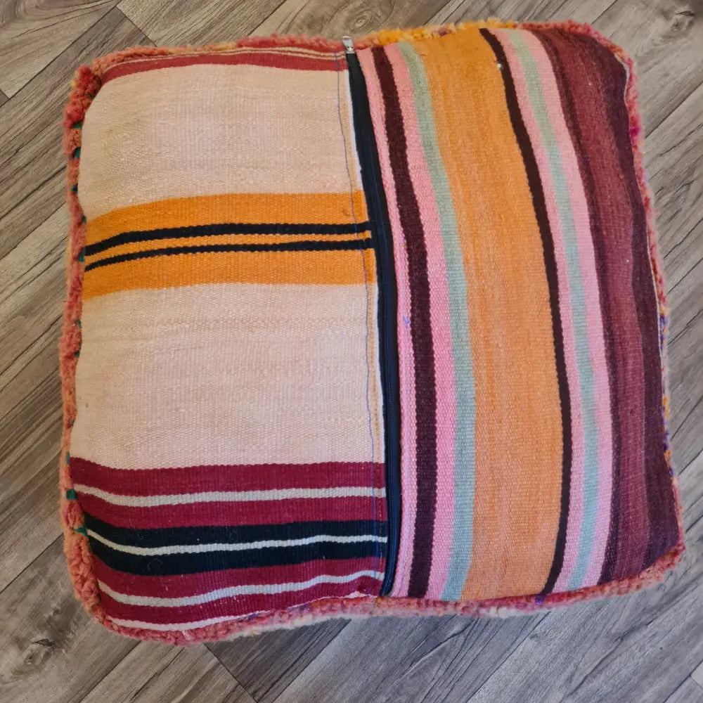 The bottom of a square Boucherouite pouf, showing a striped Kilim pattern in pink, orange, and cream, with a large black zipper for stuffing