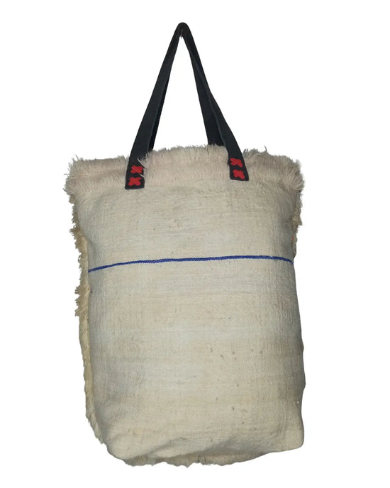 A front-view of a handmade Moroccan Berber-style tote bag on a white background. The bag is made of off-white, shaggy wool and features colorful embroidered kilim panels with geometric tribal patterns. The bag has two black leather shoulder straps and its dimensions are displayed