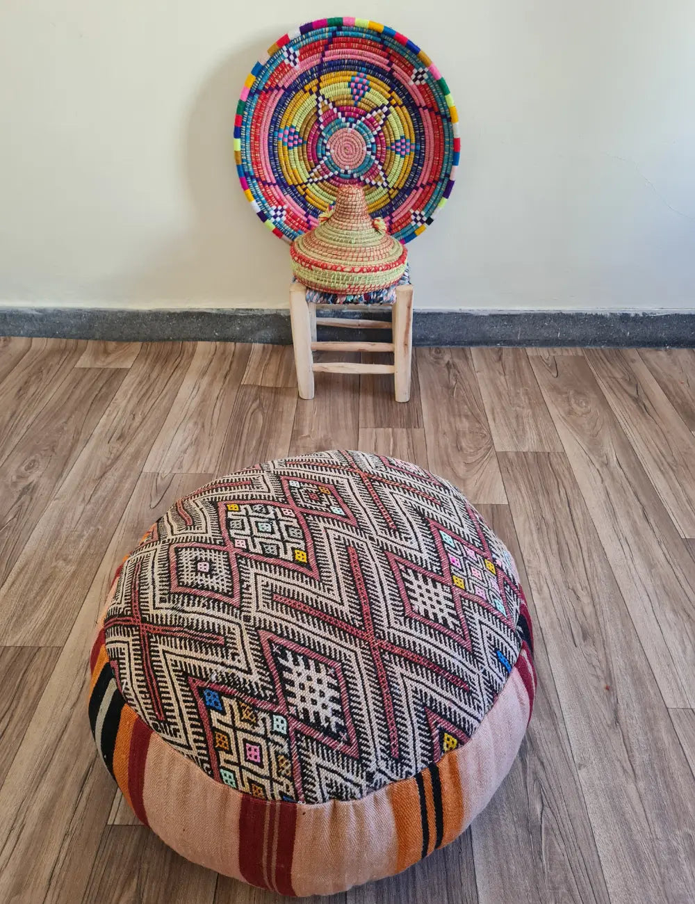 A vintage Kilim floor cushion on a wood floor. In the background is a small wooden stool with a colorful Moroccan basket and plate.