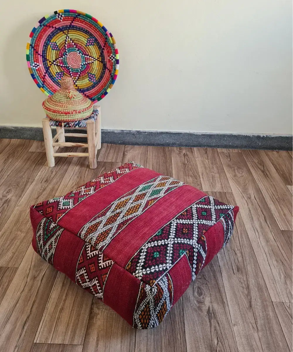 A square vintage Kilim floor cushion in rich red on a wood floor. In the background, a small wooden stool displays a colorful Moroccan woven basket