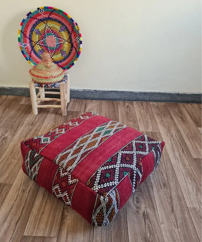 A square vintage Kilim floor cushion in rich red on a wood floor. In the background, a small wooden stool displays a colorful Moroccan woven basket