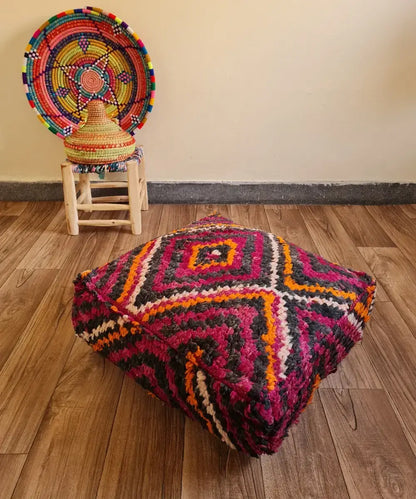 A square vintage Boucherouite floor cushion in vibrant magenta and orange on a wood floor. In the background, a small wooden stool displays a colorful Moroccan woven basket