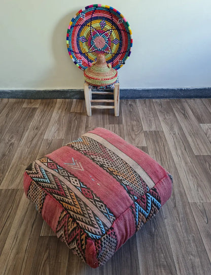 A square vintage Kilim floor cushion in shades of dusty rose on a wood floor. In the background, a small wooden stool displays a colorful Moroccan woven basket