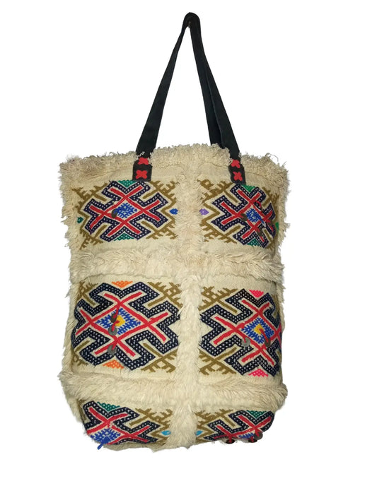 A front-view of a handmade Moroccan Berber-style tote bag on a white background. The bag is made of off-white, shaggy wool and features colorful embroidered kilim panels with geometric tribal patterns. The bag has two black leather shoulder straps and its dimensions are displayed.
