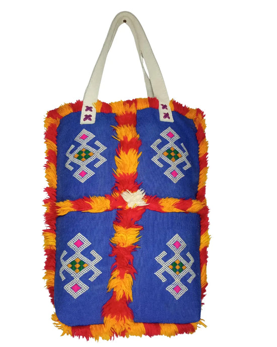 A front-view of a handmade Moroccan Berber-style tote bag on a white background. The bag is made of royal blue fabric with four panels of geometric embroidery and is trimmed with shaggy red and yellow wool fringe. It has off-white shoulder straps, and its dimensions are displayed.