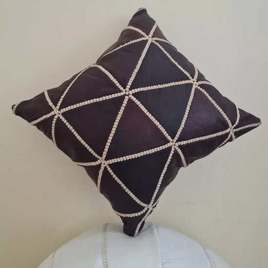 A handmade, square Moroccan throw pillow cover in dark brown leather. It features a geometric diamond pattern created with thick, off-white hand-stitching