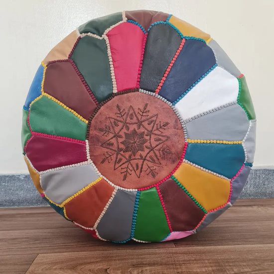 A top-down view of a round, handmade Moroccan pouf made from multi-colored leather patches. It has colorful hand-stitching and a central tan leather medallion with embroidery