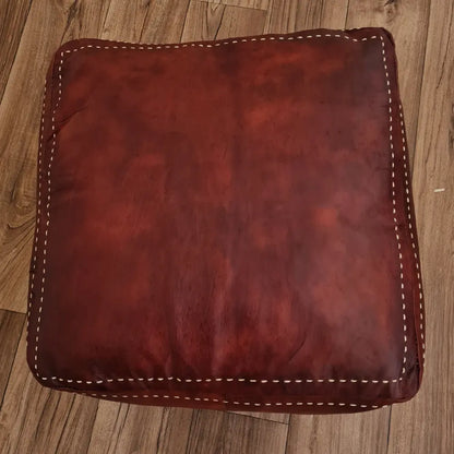 A top-down view of a handmade, square Moroccan pouf in a rich, distressed brown leather. It features thick, contrasting off-white hand-stitching along the seams