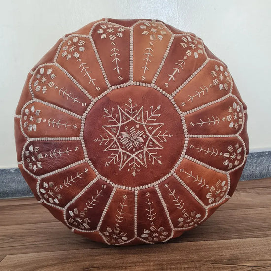 A top-down view of a classic tan, round Moroccan leather pouf. It features contrasting white hand-stitching and a central embroidered star design.