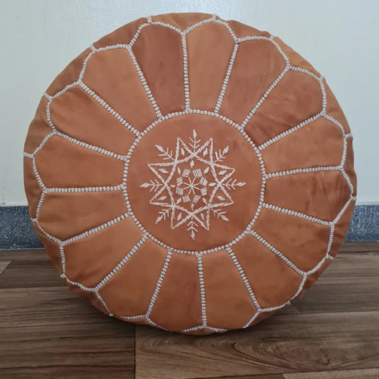 A top-down view of a round, handmade Moroccan pouf in warm tan suede. It features contrasting off-white hand-stitching and a central embroidered star design