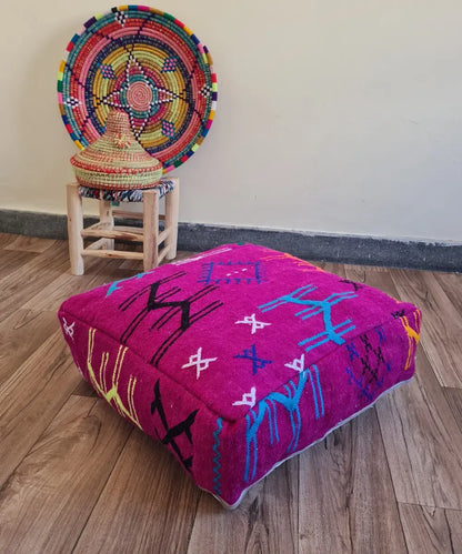 A square modern floor cushion with fuchsia embroidery on a wood floor. In the background, a small wooden stool displays a colorful Moroccan woven basket