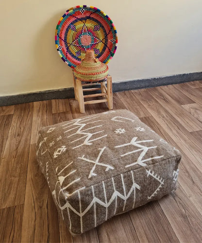 A square modern floor cushion with black and white tribal patterns on a wood floor. In the background, a small wooden stool displays a colorful Moroccan woven basket