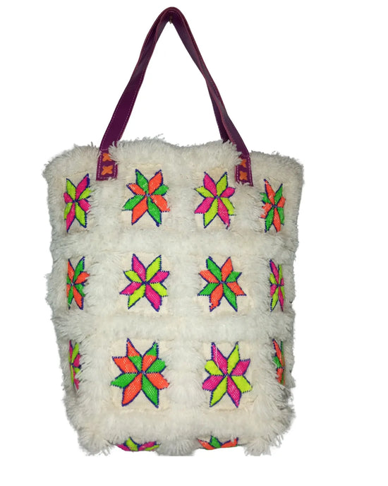A front-view of a white fuzzy sherpa tote bag on a white background. The bag is decorated with a grid of embroidered patches featuring neon starburst patterns in pink, green, and orange. It has two purple faux leather shoulder straps. Dimensions are noted on the image.