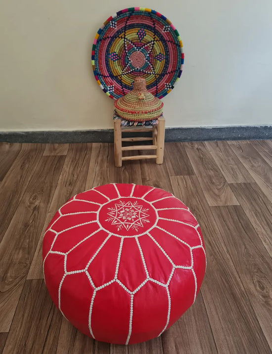 A red Moroccan leather pouf on a wood floor. In the background is a small wooden stool with a colorful Moroccan basket and plate.