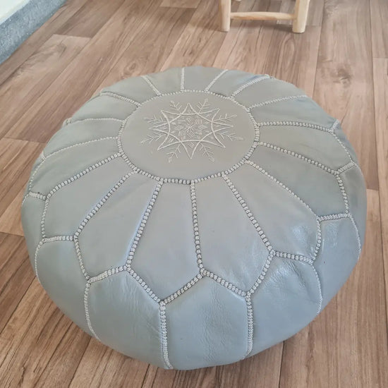 An angled, three-quarter view of a grey leather Moroccan pouf, highlighting the intricate white stitching and embroidery