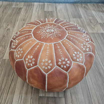 An angled, three-quarter view of a tan leather Moroccan pouf, highlighting the intricate floral embroidery on the side panels and the white stitching.