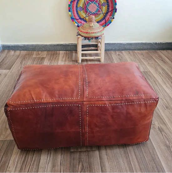 A rectangular brown leather pouf used as an ottoman bench on a wood floor. In the background is a small wooden stool with a colorful Moroccan basket
