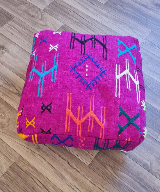 The side profile of a square modern Kilim pouf, showing the durable woven texture and the vibrant, colorful embroidery along the fuchsia side