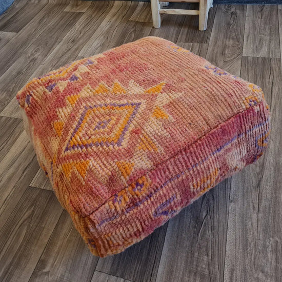 The side profile of a square vintage rug pouf, showing the beautifully worn, low-pile texture and the faded red and orange color palette